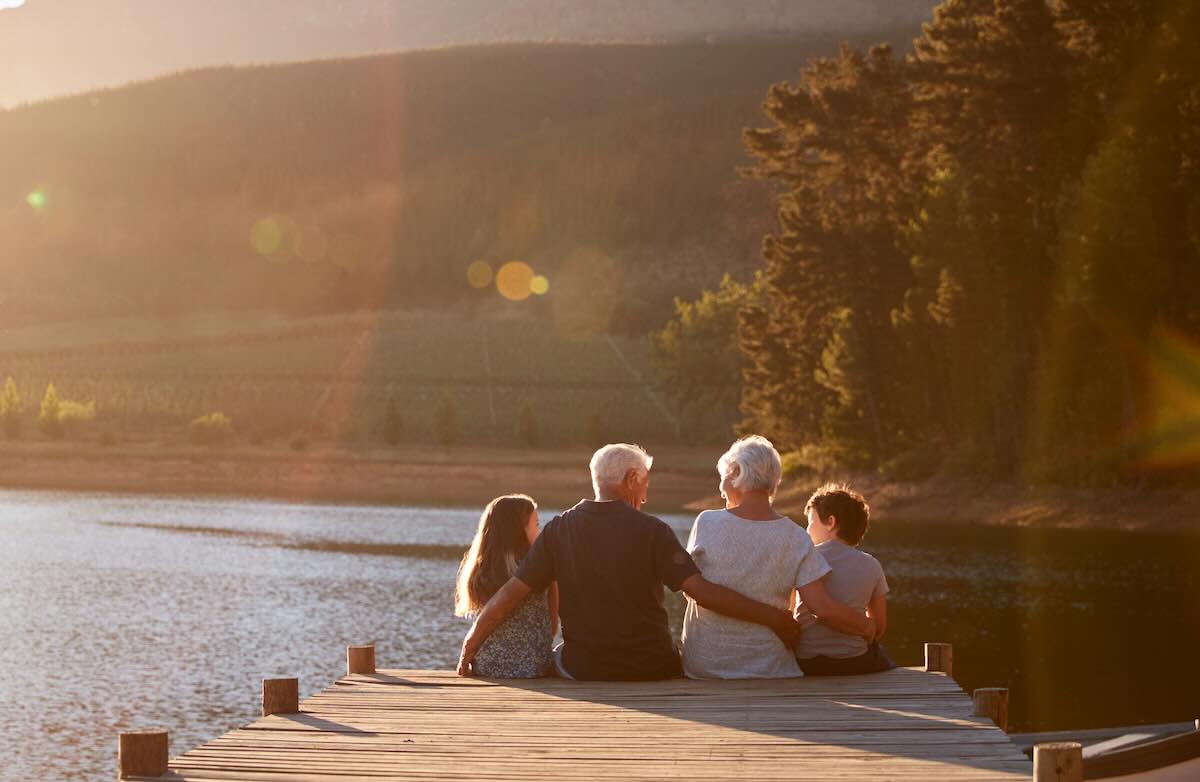 Grandchildren With Grandparents Sitting On Wooden Jetty By Lake